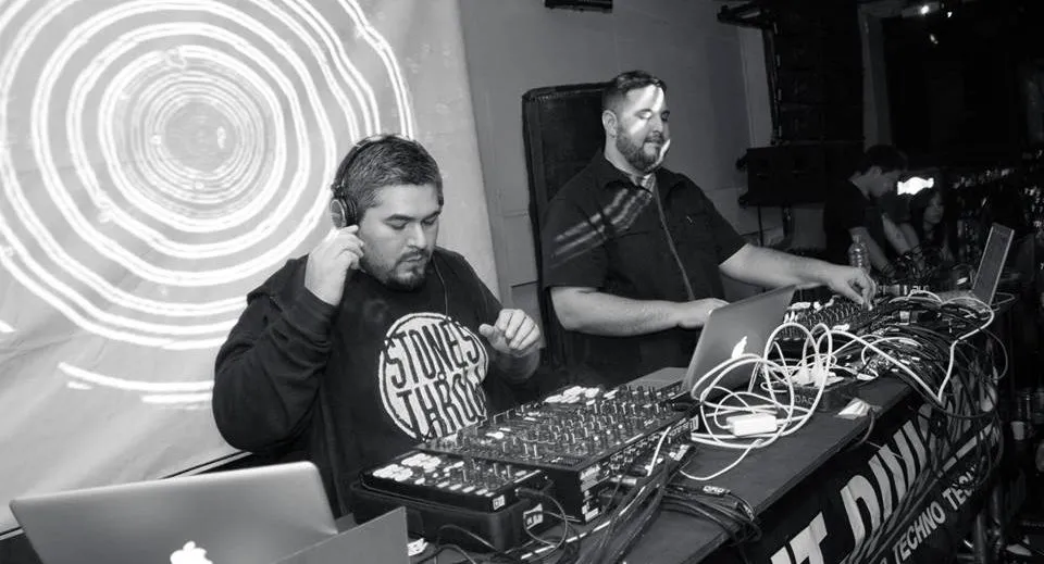 2 DJs spinning together at a table with an abstract projected background behind them